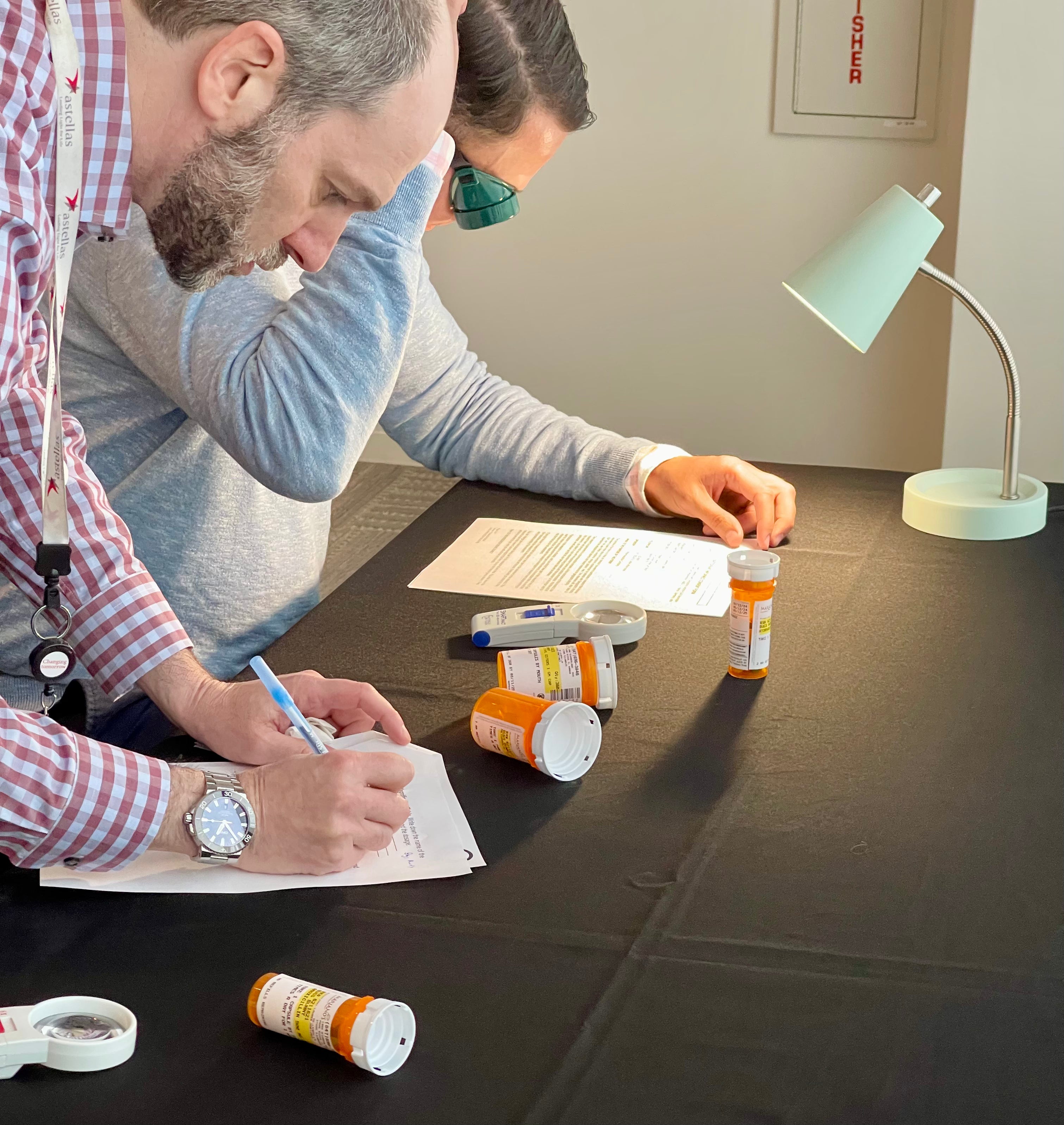 Two people , one wearing goggles, reviewing medical supplies on a table with a lamp in the background.