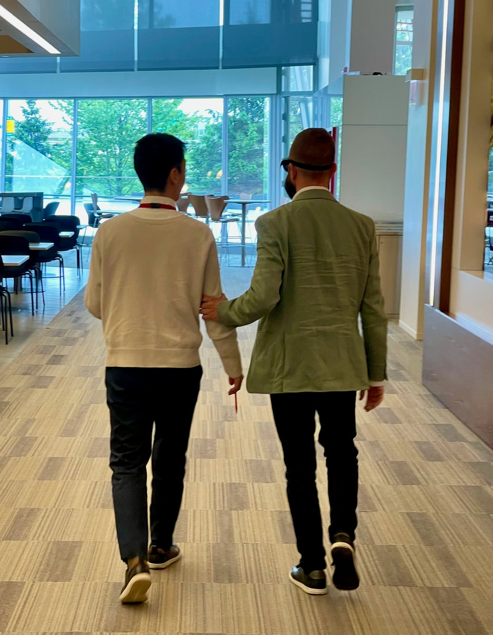 Two people walking together in a modern indoor setting using human guide and simulation goggles.