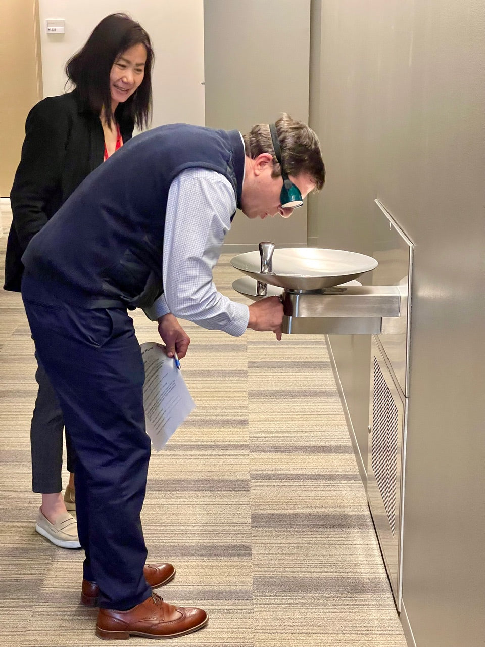 Person wearing simulation goggles using a water fountain with another person standing nearby.